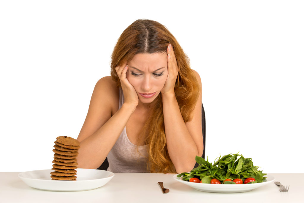 Young woman tired of diet restrictions deciding whether to eat healthy food or sweet cookies she is craving sitting at table isolated white background. Human face expression emotion. Nutrition concept